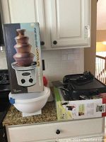 Photo showing Nostalgia stainless steel chocolate fondue fountain original boxed, stacked on white plastic food storage containers, and next to black Bella 10x16 inch griddle and George Foreman grill on kitchen counter.