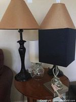 Four table lamps shown on wooden table. Two traditional black base lamps with tan conical shade, a clear glass base lamp with white shade and a modern lamp with square black shade on metal base with coiled cord.