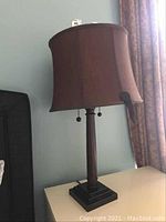 Single lamp with brown fabric shade and dark wood base shown on white surface near window with curtains.