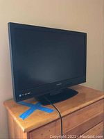 Full view of the black framed 32-inch Sylvania LCD television on a wooden dresser, showing the screen, base stand, and power cable.