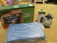 Photo showing boxed Coleman Perfectflow 2-burner stove, sealed new, beside a boxed LED hurricane lamp and a blue hard case.
