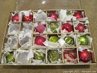 24 hand painted glass Christmas ornaments in original box with white tissue separators. Various colors and designs visible including red, green, white glitter, and clear with snowflake patterns.