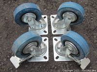 Top-down view of four blue rubber casters on black surface. Two casters have visible brake locks.