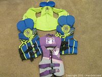 All four youth life jackets shown together on floor; three blue and yellow Hyperlite jackets and one purple, pink and grey Bodyglove jacket.