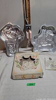 Two aluminum cake pans in novelty shapes and boxed ceramic shortbread pan with recipe booklet visible on table