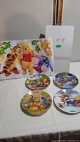 Front view of the 4 Disney Winnie the Pooh collector plates with seasonal character illustrations and a large Pooh and Tigger sign behind.
