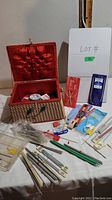 Wicker sewing box open showing red fabric interior and many knitting needles and accessories spread around it.