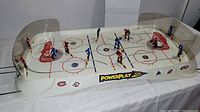 Full view of Power Play 2 table hockey game showing player figures on rods in red and blue, hockey rink surface with logos, and goals; plastic puck shields are visible and damaged.
