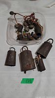 Three vintage camel bells of varying sizes on white fabric surface next to a plastic container with smaller bells and string with beads.