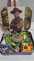 Open tin box filled with colorful vintage Boy Scout patches including a large Wolf Cubs patch, Cub Fun 1980, camping themed patch, and others.