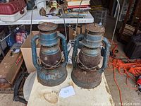 Two blue metal Dietz Little Wizard barn lanterns on a table showing surface rust and dirt.