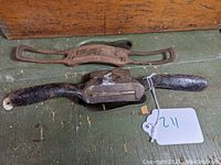 Top view of two vintage metal spokeshaves on a green wooden surface with a brown wooden box lid behind. One spokeshave has a rusted full metal frame and the other has black wooden handles.