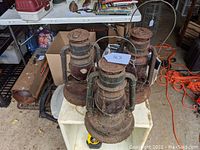 Three rusted metal Dietz Little Giant lanterns with red glass globes shown on a white shelf indoors. Wires, orange extension cord, and other items in background.