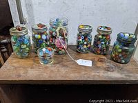 Seven glass jars filled with colorful marbles, arranged in one row on a wooden surface.