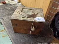 Wooden rectangular box with leather handle and metal knob on front, shown on workbench surface.