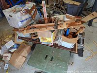 Photo showing wooden bench with boxes containing hand saws, saw blades, and miscellaneous tools atop it, along with a green folding wooden table in front.
