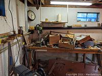View of the wooden workbench with assorted hand tools, cords, boxes with parts, and various small containers. Also shows drill press near bench (not included).