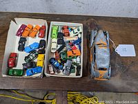 Top down view showing two trays of multiple colorful die-cast toy cars and a large orange and silver metal toy car.