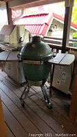 Full view of green Big Green Egg grill on metal rolling stand with wooden side tables on deck
