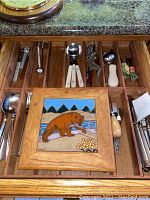 Wooden kitchen drawer containing stainless steel silverware and various kitchen utensils and tools, including measuring spoons, bottle openers, spatula, whisk, and a decorative bear tile.
