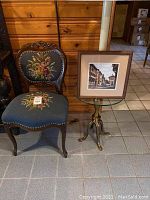 Full view of the antique Victorian style needlepoint chair with floral upholstery, the round glass top lamp table with brass base holding the framed cityscape picture.