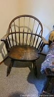 Front view of vintage wooden Windsor sack back rocking chair with spindle back and armrests, showing wear and dark wood finish.