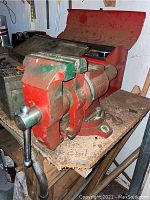 Photo showing the red and green heavy-duty bench vise mounted on a wooden workbench with visible wear and dirt