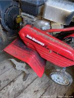 Close-up of red Yard Machines rototiller with metal tines and small wheels, showing wear and dirt.