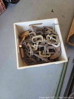 Overview of white plastic crate filled with metal C-clamps of various sizes and types, some having wooden handles.