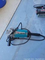 Photo of a blue Makita electric disc grinder with side handle and power cord, tool resting on a concrete floor surface.
