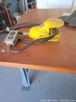 Yellow Square D foot switch placed on a wooden table showing power cord and attached metal outlet box with two electrical outlets.
