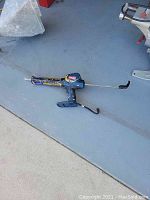 Photo showing a used blue Ryobi cordless grout removal tool lying on a floor with its grout scraping attachment, and a connected 18V battery pack.