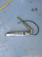 Hand operated metal grease gun with a lever and flexible black hose nozzle lying on a concrete floor.