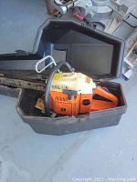 Husqvarna Rancher chainsaw placed in an open black hard carrying case on a grey concrete floor, with a miter saw partially visible in the background.