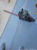 Photo showing used gas-powered hedge trimmer lying on a concrete floor, revealing the engine unit, handles, and long cutting blades with a blade guard.