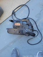 Black Craftsman electric belt sander laying on gray floor, showing main body, handle, and attached cord.