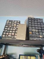 Three storage cases filled with assorted screws, bolts, nuts, washers, and other small hardware items positioned on a metal shelf surface against a wall.