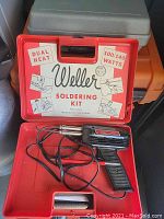 Red plastic case with Weller branding and dual heat soldering kit label. Contains a pistol-style soldering iron and metal stand.