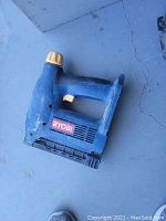 Blue Ryobi cordless stapler on gray concrete surface showing front and side profile
