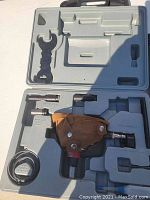 Top-down photo of open molded plastic carry case revealing steel finish nailer with leather holster, various metal parts, adapter with tubing, and wrench tool.