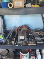 Photo showing shelves with multiple socket sets, analog meter, coiled copper wire, welding rods, and various small metal tools.