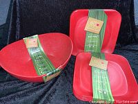 Full view of one red bowl and a stack of eight matching square plates, all with EcoSmart poly-flax labels attached. Set displayed on black fabric background.
