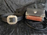 Crossbody bag and belt displayed on black textured fabric. Bag is croc embossed two tone with heart decoration. Belt is black with large silver-tone engraved western buckle.