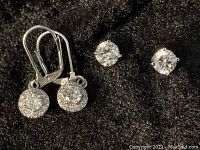 Two pairs of simulated diamond earrings on black fabric - one pair lever back with halo setting and one pair simple stud earrings.