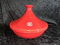 Red Emile Henry ceramic tagine with cone-shaped lid marked with EH logo, displayed on black background, showing full side view.