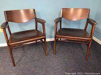 Pair of vintage walnut wooden chairs with cushioned brown seats and wooden armrests, placed on carpeted floor against blue wall.
