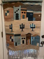 Full view of the wool wall hanging with desert village scene and cacti, including fringed bottom and wooden hanger.
