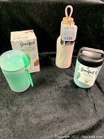Photo showing the trio of food storage items on a black background: the mint green Goodful snack container with a box, cream-colored Primula insulated bottle with wood top, and gray-lidded Goodful food jar.
