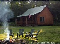 Exterior view of wood cabin with green metal roof, covered porch, Adirondack chairs and fire pit in clearing