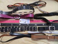 Eleven assorted belts laid out on a cream fabric background showing varied styles including braided, grommet, and solid buckled belts. Colors include black, brown, purple, white, and tan.